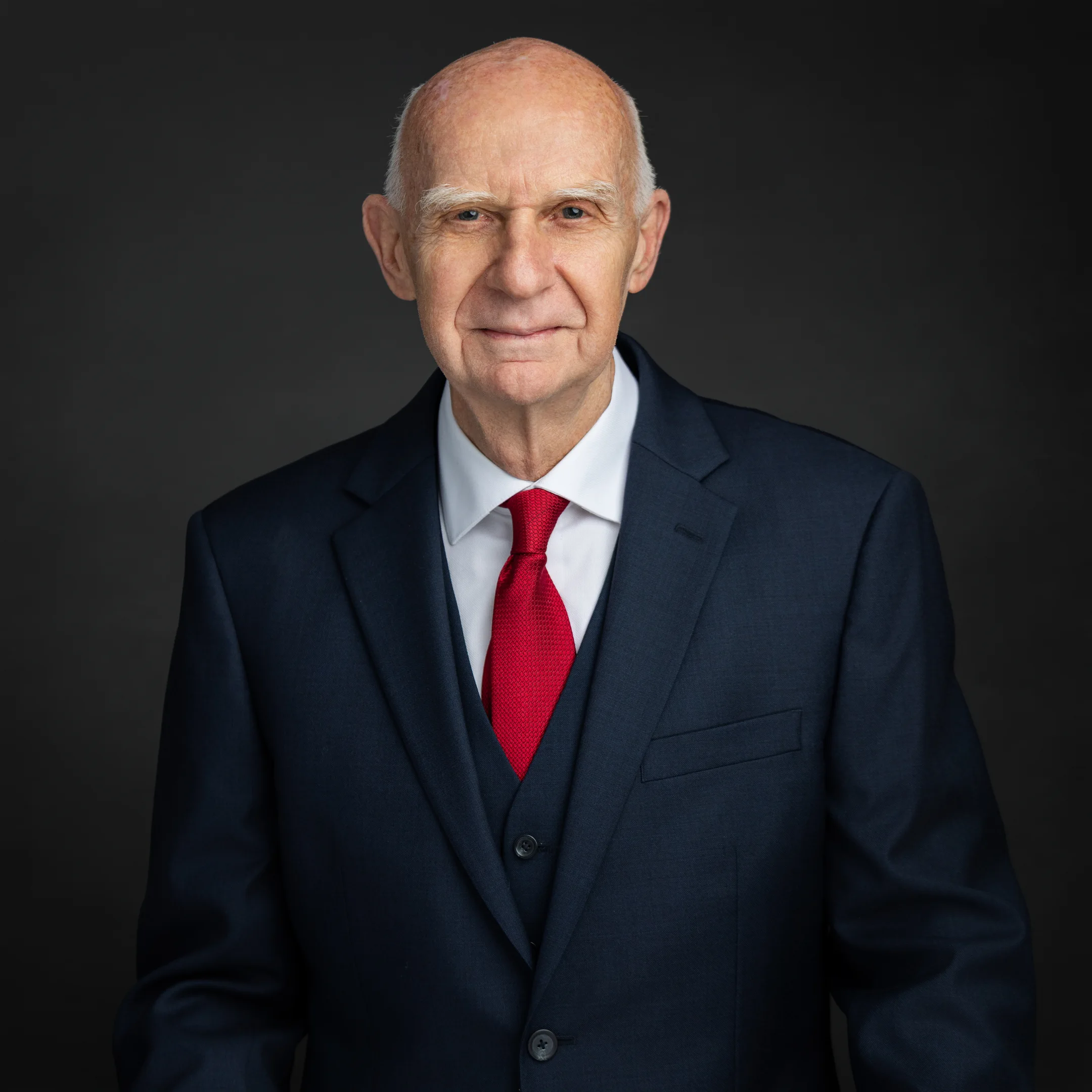 An older man with short gray hair in a blue suit, white shirt, and red tie poses on a dark background for Delray Beach product photography.