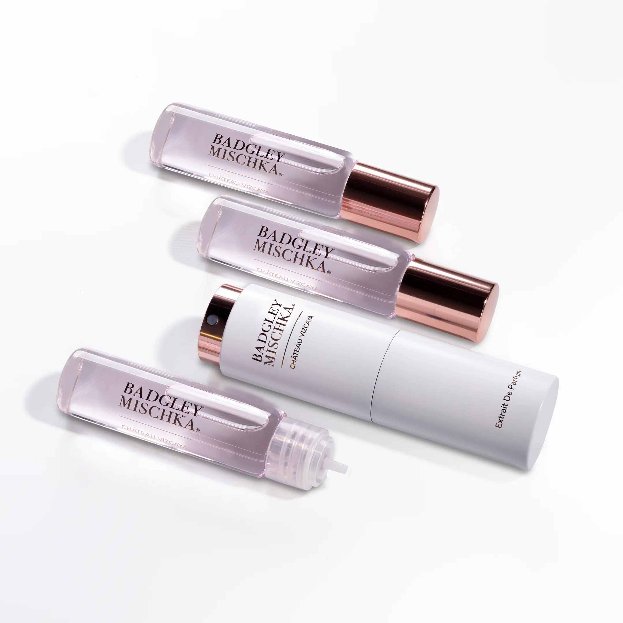 Clear glass perfume bottles with rose gold caps and a BADGLEY MISCHKA spray sit on white, expertly shot for South Florida brands.